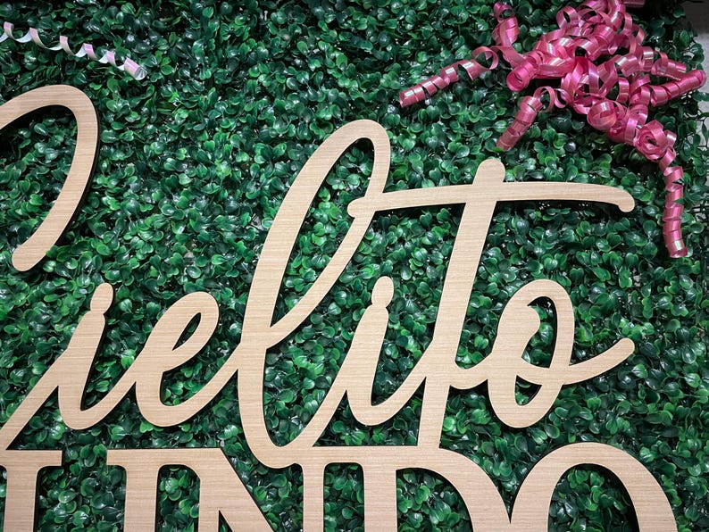 May include: Wooden sign with the words "Cielito Lindo" in a cursive font. The sign is made of light-colored wood and is set against a backdrop of green foliage. A pink ribbon is in the upper right corner.