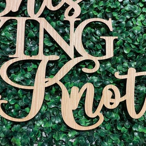 Puede incluir: Letrero de madera con las palabras "Tying the Knot" en fuente cursiva. El letrero est&aacute; hecho de madera de color claro y est&aacute; colocado sobre un fondo de follaje verde. Ideal para la decoraci&oacute;n de bodas.