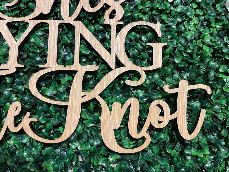 Puede incluir: Letrero de madera con las palabras "Tying the Knot" en fuente cursiva. El letrero est&aacute; hecho de madera de color claro y est&aacute; colocado sobre un fondo de follaje verde. Ideal para la decoraci&oacute;n de bodas.