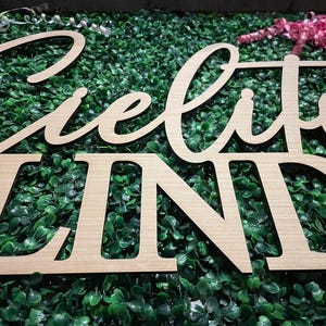 May include: Wooden sign spelling "Cielito Lindo" in a cursive and block font. The sign is made of light-colored wood and is set against a backdrop of green foliage. Decorative ribbons are visible in the background.