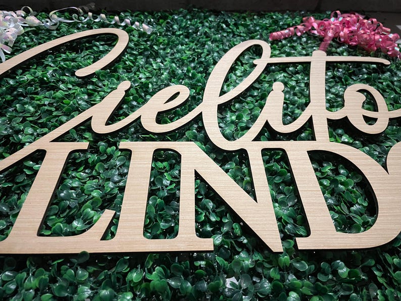 May include: Wooden sign spelling "Cielito Lindo" in a cursive and block font. The sign is made of light-colored wood and is set against a backdrop of green foliage. Decorative ribbons are visible in the background.