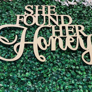 Peut inclure: Panneau en bois avec l'inscription "SHE FOUND HER Honey" dans une police cursive. Le panneau est en bois de couleur claire et est plac&eacute; sur un fond de feuillage vert. Des rubans d&eacute;coratifs sont visibles en arri&egrave;re-plan.