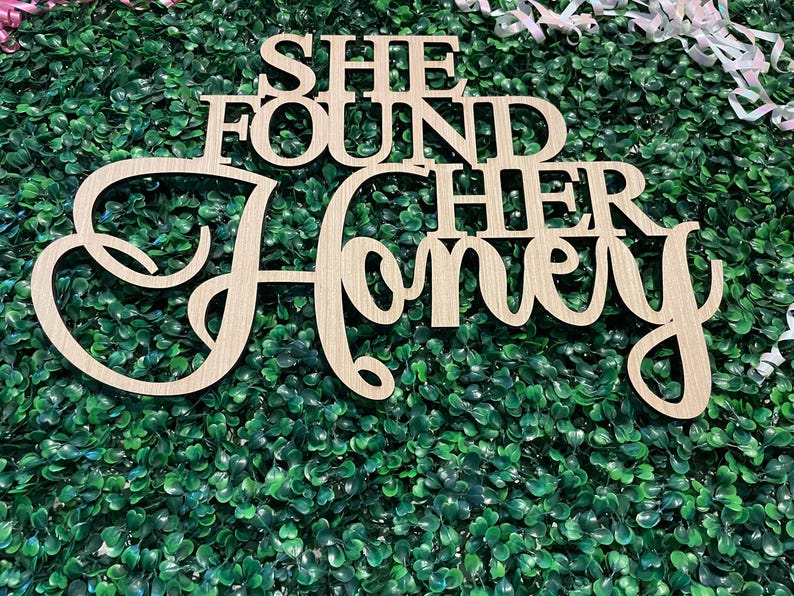 Peut inclure: Panneau en bois avec l'inscription "SHE FOUND HER Honey" dans une police cursive. Le panneau est en bois de couleur claire et est plac&eacute; sur un fond de feuillage vert. Des rubans d&eacute;coratifs sont visibles en arri&egrave;re-plan.