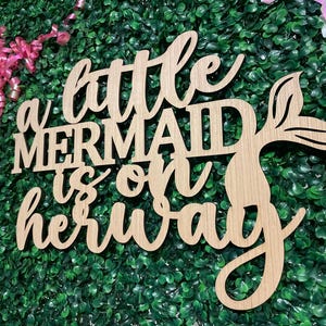 May include: Wooden sign with the phrase "a little mermaid is on her way" in cursive and block letters. The sign features a mermaid tail design. The background is a green hedge with pink and white ribbons.