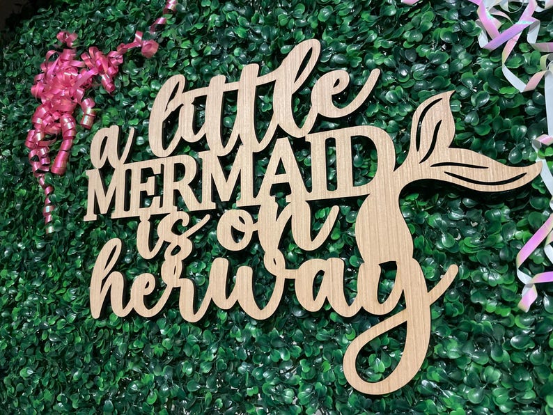 May include: Wooden sign with the phrase "a little mermaid is on her way" in cursive and block letters. The sign features a mermaid tail design. The background is a green hedge with pink and white ribbons.