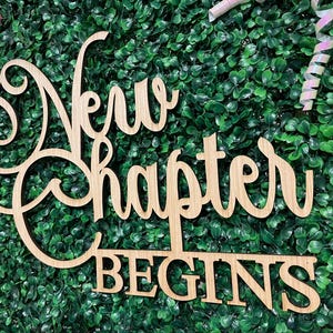 May include: Wooden sign with the words "A New Chapter Begins" in a decorative font. The sign is made of light-colored wood and is set against a backdrop of green foliage. The sign is a decorative item.