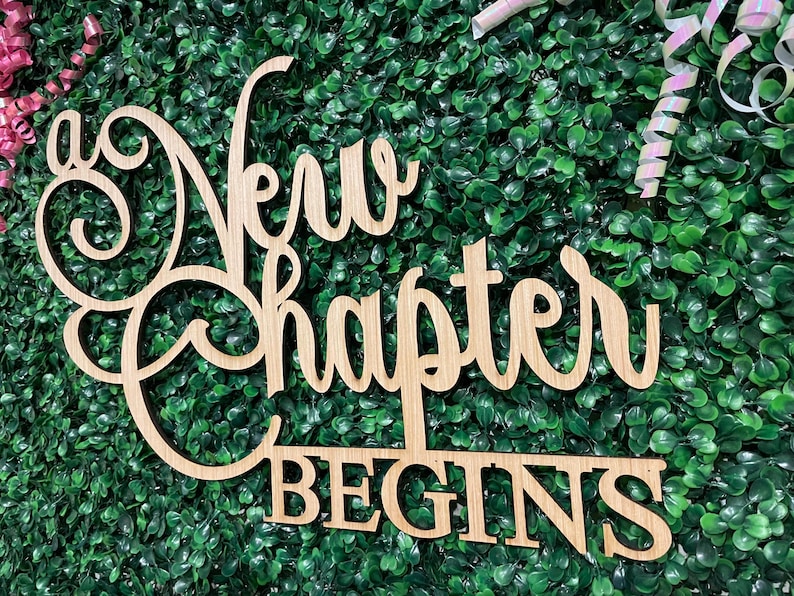 May include: Wooden sign with the words "A New Chapter Begins" in a decorative font. The sign is made of light-colored wood and is set against a backdrop of green foliage. The sign is a decorative item.