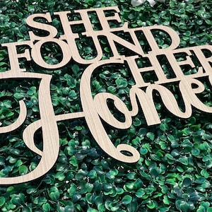 Peut inclure: Panneau en bois clair d&eacute;coup&eacute; avec l'inscription "SHE FOUND HER Honey" en police cursive. Le panneau est pos&eacute; sur un fond de feuillage vert, avec des rubans d&eacute;coratifs en arri&egrave;re-plan.