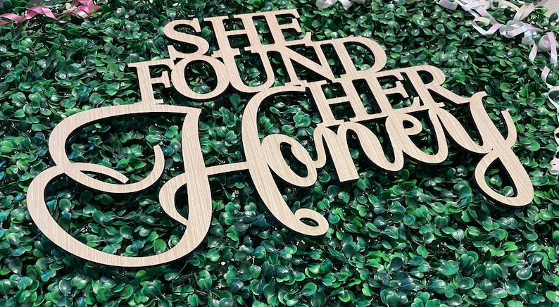 Peut inclure: Panneau en bois clair d&eacute;coup&eacute; avec l'inscription "SHE FOUND HER Honey" en police cursive. Le panneau est pos&eacute; sur un fond de feuillage vert, avec des rubans d&eacute;coratifs en arri&egrave;re-plan.