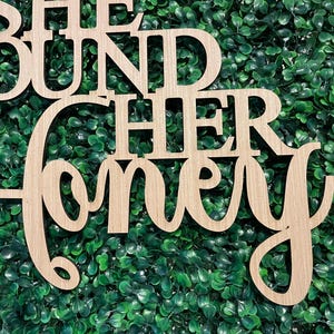 Peut inclure: Panneau en bois avec la phrase "SHE FOUND HER HONEY" d&eacute;coup&eacute;e dans une police d&eacute;corative. Le panneau est en bois de couleur claire et est plac&eacute; sur un fond de feuillage vert, avec quelques rubans dans le coin sup&eacute;rieur droit.