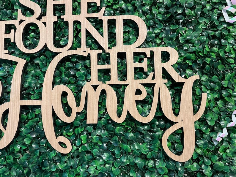 Peut inclure: Panneau en bois avec la phrase "SHE FOUND HER HONEY" d&eacute;coup&eacute;e dans une police d&eacute;corative. Le panneau est en bois de couleur claire et est plac&eacute; sur un fond de feuillage vert, avec quelques rubans dans le coin sup&eacute;rieur droit.