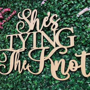 Puede incluir: Letrero de madera con la frase "She's Tying The Knot" en fuente cursiva. El letrero est&aacute; hecho de madera de color claro y est&aacute; colocado sobre un fondo de follaje verde. Rizos de cinta rosa en la esquina superior izquierda.