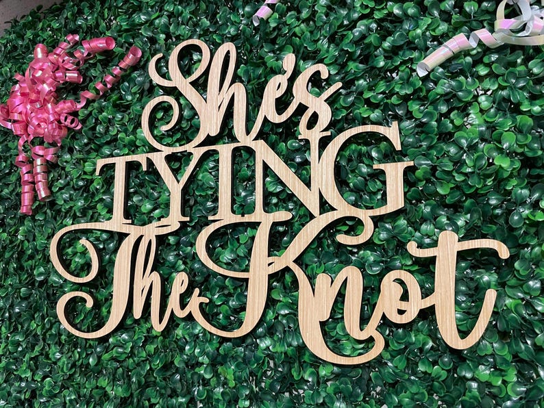 Puede incluir: Letrero de madera con la frase "She's Tying The Knot" en fuente cursiva. El letrero est&aacute; hecho de madera de color claro y est&aacute; colocado sobre un fondo de follaje verde. Rizos de cinta rosa en la esquina superior izquierda.