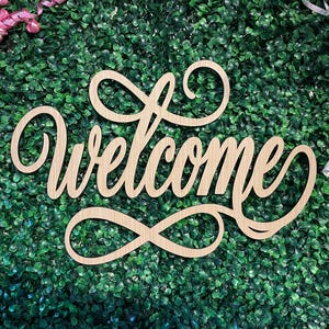 May include: A light-colored wooden cutout sign featuring the word Welcome in a cursive script font, accented with an infinity symbol at the bottom and decorative swirls at the top, displayed against a background of green artificial foliage.