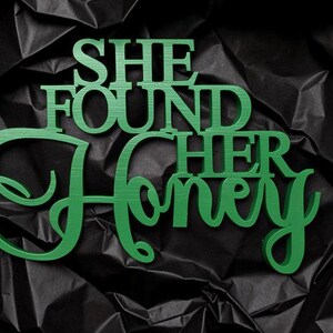 K&ouml;nnte beinhalten: Ein gr&uuml;nes Holzschild mit dem Schriftzug "SHE FOUND HER HONEY" in einer Kombination aus Block- und Schreibschrift. Das Schild hat eine glatte, matte Oberfl&auml;che und ist vor einem dunklen, zerknitterten, schwarzen Hintergrund platziert.