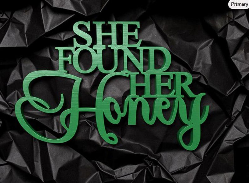 K&ouml;nnte beinhalten: Ein gr&uuml;nes Holzschild mit dem Schriftzug "SHE FOUND HER HONEY" in einer Kombination aus Block- und Schreibschrift. Das Schild hat eine glatte, matte Oberfl&auml;che und ist vor einem dunklen, zerknitterten, schwarzen Hintergrund platziert.
