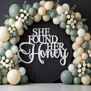 K&ouml;nnte beinhalten: Ein runder schwarzer Holzhintergrund mit dem wei&szlig;en Schriftzug SHE FOUND HER HONEY, umgeben von einem Bogen aus salbeigr&uuml;nen, cremefarbenen und wei&szlig;en Luftballons, durchsetzt mit kleinen wei&szlig;en Rosen und gr&uuml;nen Eukalyptusbl&auml;ttern.