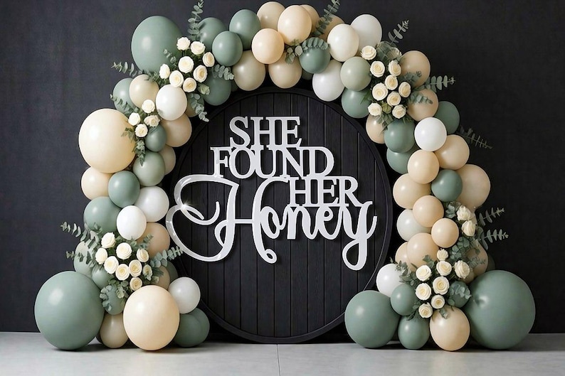 K&ouml;nnte beinhalten: Ein runder schwarzer Holzhintergrund mit dem wei&szlig;en Schriftzug SHE FOUND HER HONEY, umgeben von einem Bogen aus salbeigr&uuml;nen, cremefarbenen und wei&szlig;en Luftballons, durchsetzt mit kleinen wei&szlig;en Rosen und gr&uuml;nen Eukalyptusbl&auml;ttern.