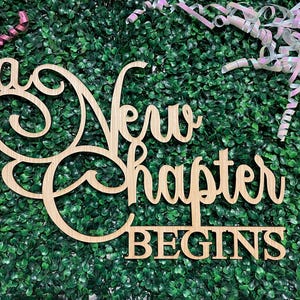 May include: A wooden sign with the words "A New Chapter Begins" in a decorative font. The sign is light brown and set against a green background with pink and white ribbon accents.