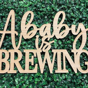 May include: A wooden sign with the text "A baby is brewing" in a cursive font. The sign is light brown and has a natural wood grain.