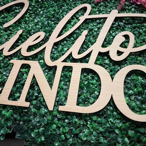 May include: Wooden sign with the words "Cielito Lindo" in a cursive font and block letters. The sign is made of light-colored wood and is set against a backdrop of green foliage. Decorative pink ribbons are visible in the background.