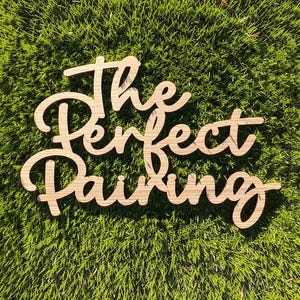 May include: A light brown wooden sign featuring the laser-cut text The Perfect Pairing in a cursive script font. The sign consists of three stacked lines of connected letters with a visible wood grain texture throughout the entire surface.
