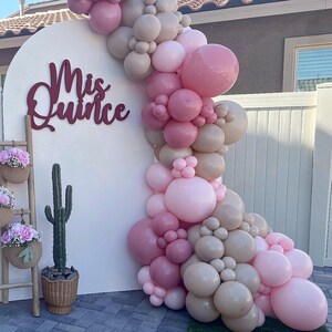Op de afbeelding: Een witte boogvormige achtergrond met de bordeauxrode tekst Mis Quince, versierd met een grote ballonslinger in roze en beige tinten. Een houten ladder bevat twee gevlochten manden met roze bloemen en een cactus in een pot staat op de grond.