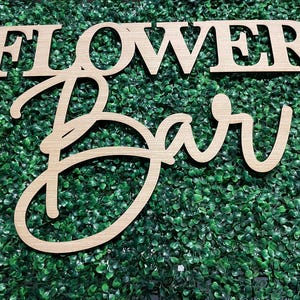 May include: Wooden sign with the words "Flower Bar" in a cursive font. The sign is made of light brown wood and is set against a backdrop of green artificial foliage.