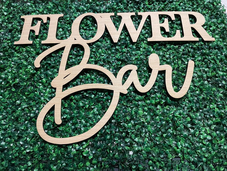 May include: Wooden sign with the words "Flower Bar" in a cursive font. The sign is made of light brown wood and is set against a backdrop of green artificial foliage.