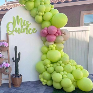 Op de afbeelding: Een witte boogvormige achtergrond met de groene tekst Mis Quince, versierd met een grote ballonslinger in limoengroen en roze. Een houten ladder bevat drie gevlochten manden met roze bloemen en er staat een cactus in een pot naast.