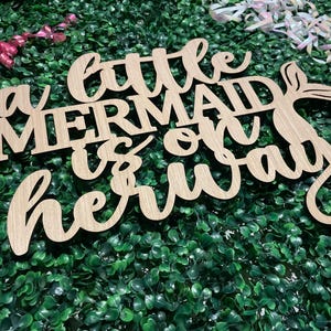 May include: Wooden sign with the text "a little mermaid is on her way" in cursive and block letters. The sign has a mermaid tail design. The sign is on a green background with pink and white ribbons.
