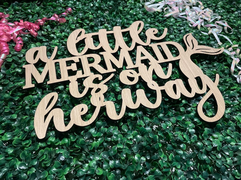 May include: Wooden sign with the text "a little mermaid is on her way" in cursive and block letters. The sign has a mermaid tail design. The sign is on a green background with pink and white ribbons.