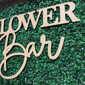 May include: Wooden sign with the words "Flower Bar" in a cursive font, set against a backdrop of green artificial foliage.