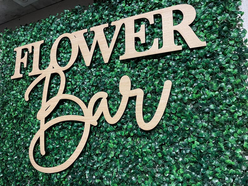 May include: Wooden sign with the words "Flower Bar" in a cursive font, set against a backdrop of green artificial foliage.