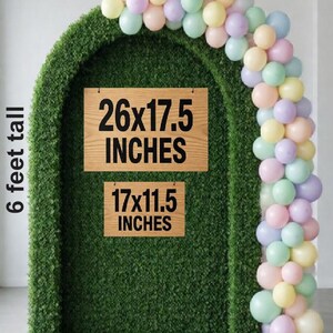 Op de afbeelding: Een groene boogvormige achtergrond met een pastelkleurige ballonslinger aan de rechterkant. Twee rechthoekige houten borden worden getoond, &eacute;&eacute;n met het label 26x17.5 INCHES en de andere 17x11.5 INCHES. Bovenaan staat SIGN CHOICES SIZE CHART.