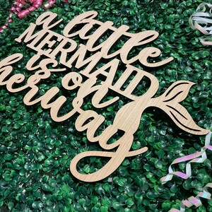 May include: Wooden sign with the phrase "a little mermaid is on her way" in a cursive font. The sign includes a mermaid tail design. The sign is light brown and rests on a bed of green foliage, with pink and white ribbons.