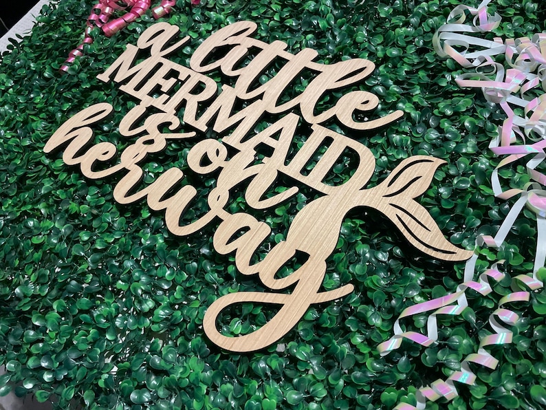 May include: Wooden sign with the phrase "a little mermaid is on her way" in a cursive font. The sign includes a mermaid tail design. The sign is light brown and rests on a bed of green foliage, with pink and white ribbons.