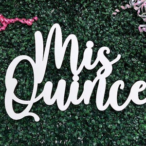 May include: A white cutout sign featuring the cursive text Mis Quince, positioned against a dark green artificial foliage background, accented with pink curling ribbon on the top left and iridescent white curling ribbon on the top right.