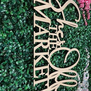 Puede incluir: Letrero de madera con la frase "She's Off the Market" en letras cursivas y de bloque. El letrero es de madera clara y est&aacute; sobre un fondo de follaje verde. Tambi&eacute;n se ven cintas rosas y blancas.