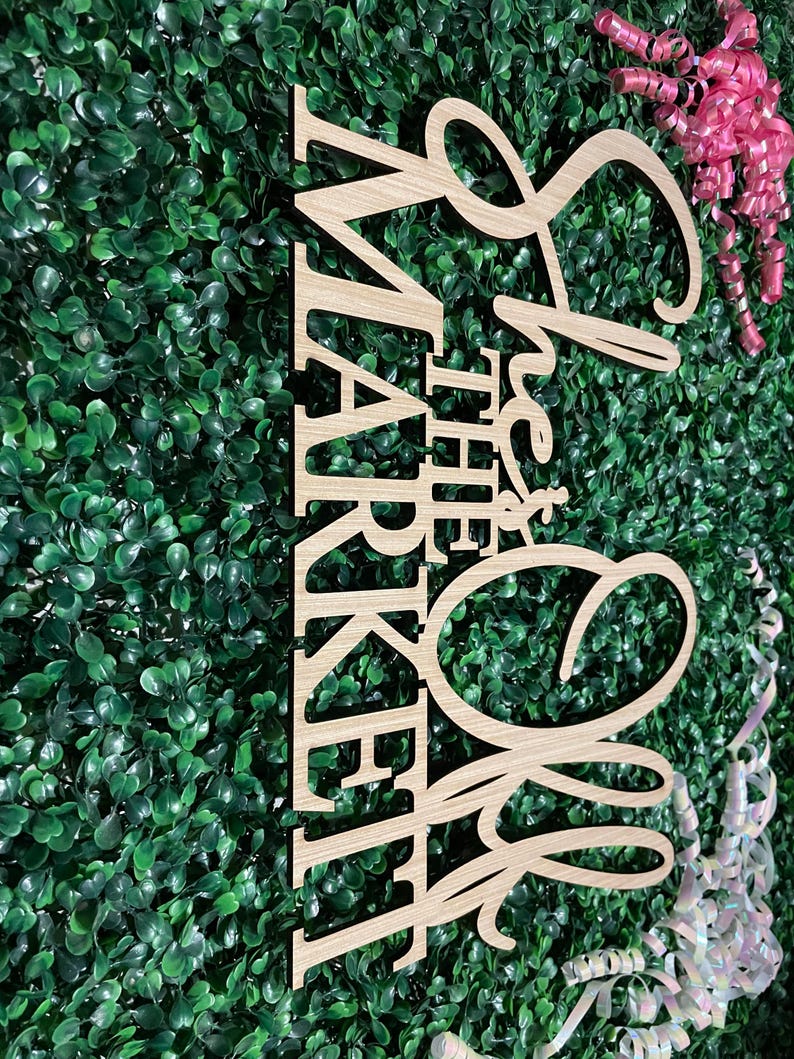 Puede incluir: Letrero de madera con la frase "She's Off the Market" en letras cursivas y de bloque. El letrero es de madera clara y est&aacute; sobre un fondo de follaje verde. Tambi&eacute;n se ven cintas rosas y blancas.