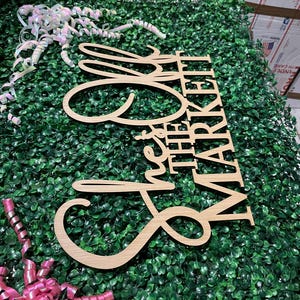 Puede incluir: Un letrero de madera que dice "She's the Market" en una fuente cursiva. El letrero est&aacute; hecho de madera de color claro y se coloca sobre un lecho de follaje artificial verde. Cintas rosas y blancas est&aacute;n esparcidas alrededor del letrero.