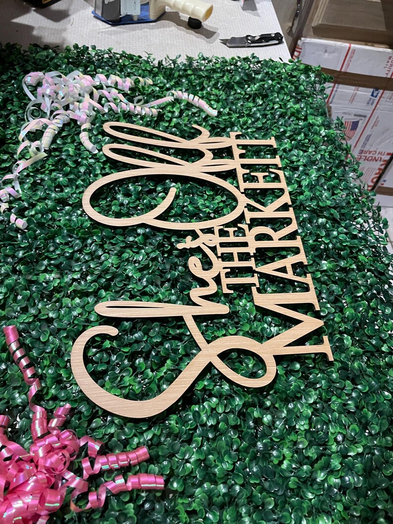 Puede incluir: Un letrero de madera que dice "She's the Market" en una fuente cursiva. El letrero est&aacute; hecho de madera de color claro y se coloca sobre un lecho de follaje artificial verde. Cintas rosas y blancas est&aacute;n esparcidas alrededor del letrero.