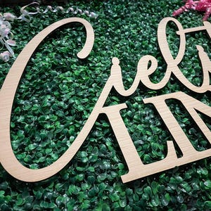 May include: Wooden sign spelling out "Cielito Lindo" in a cursive font. The sign is made of light-colored wood and is set against a backdrop of green foliage. Decorative ribbons are visible in the background.