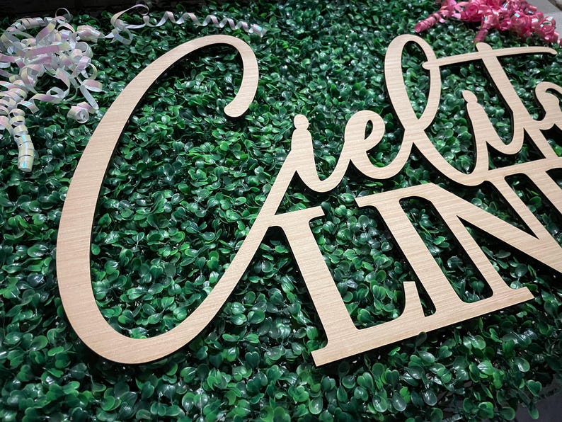 May include: Wooden sign spelling out "Cielito Lindo" in a cursive font. The sign is made of light-colored wood and is set against a backdrop of green foliage. Decorative ribbons are visible in the background.