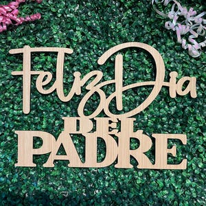 May include: A light-colored wooden cutout sign featuring the Spanish text Feliz Dia del Padre in a combination of cursive and block lettering. The sign is positioned against a background of green artificial foliage with pink and iridescent ribbons.