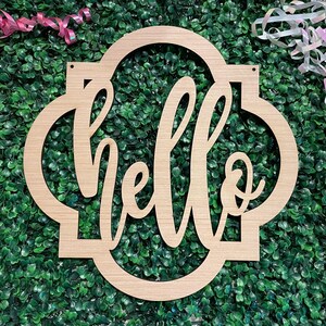 May include: A wooden sign with the word "hello" in a cursive font, set within a decorative frame. The sign is made of light-colored wood and has two small holes at the top for hanging. Pink and white ribbons are in the background.