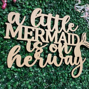 May include: Wooden sign with the phrase "a little mermaid is on her way" in cursive and block letters. The sign has a mermaid tail design and is set against a green backdrop with pink and white streamers.