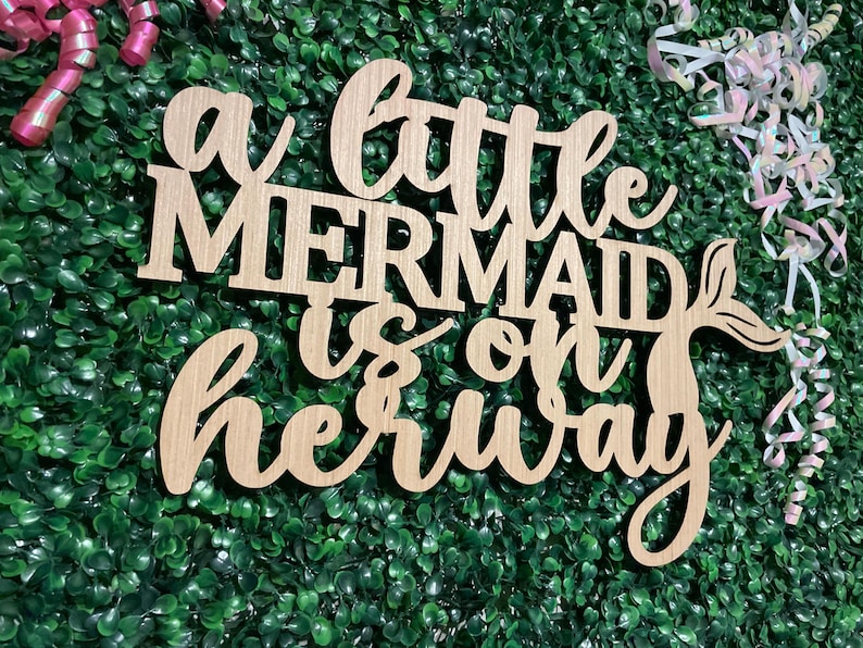 May include: Wooden sign with the phrase "a little mermaid is on her way" in cursive and block letters. The sign has a mermaid tail design and is set against a green backdrop with pink and white streamers.