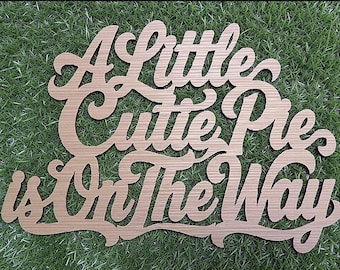May include: A light brown wooden cutout sign featuring the cursive text A Little Cutie Pie is On The Way. The letters are connected in a single piece with a smooth finish and a visible wood grain texture throughout the entire surface.