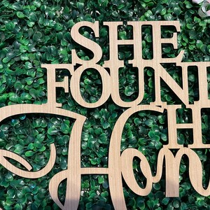 Peut inclure: Panneau en bois avec l'inscription "SHE FOUND HER HOME" dans une police d&eacute;corative. Le panneau est en bois de couleur claire et est plac&eacute; sur un fond de feuillage vert.